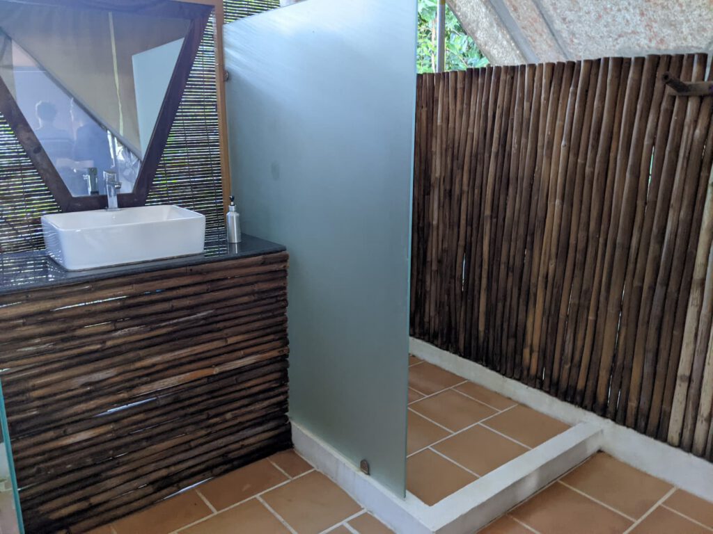 A wooden bathroom vanity is on the left with a white square basin. On the right is an open shower with one side glass screen. The walls are made from bamboo and you can see the trees outside the top of the wall.