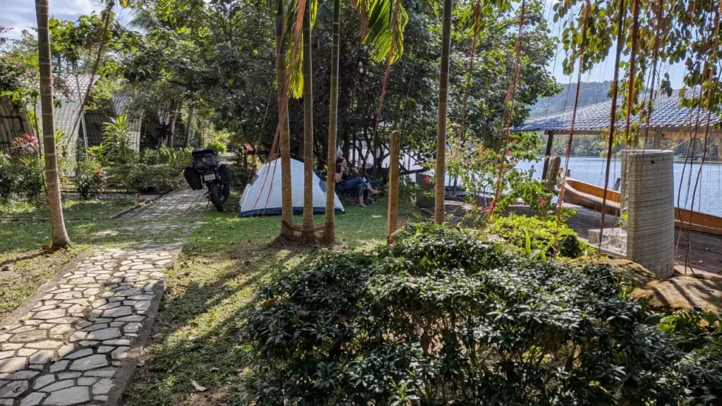 In a garden next to the Tatai river is a tent next to a garden path. A person sits in front of the tent looking at the river and a motorbike is parked behind.