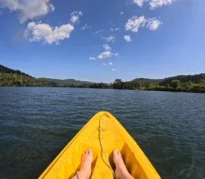 The front of a yellow kayak with two feet in it points towards the river with a blue sky and green forest surrounding it.