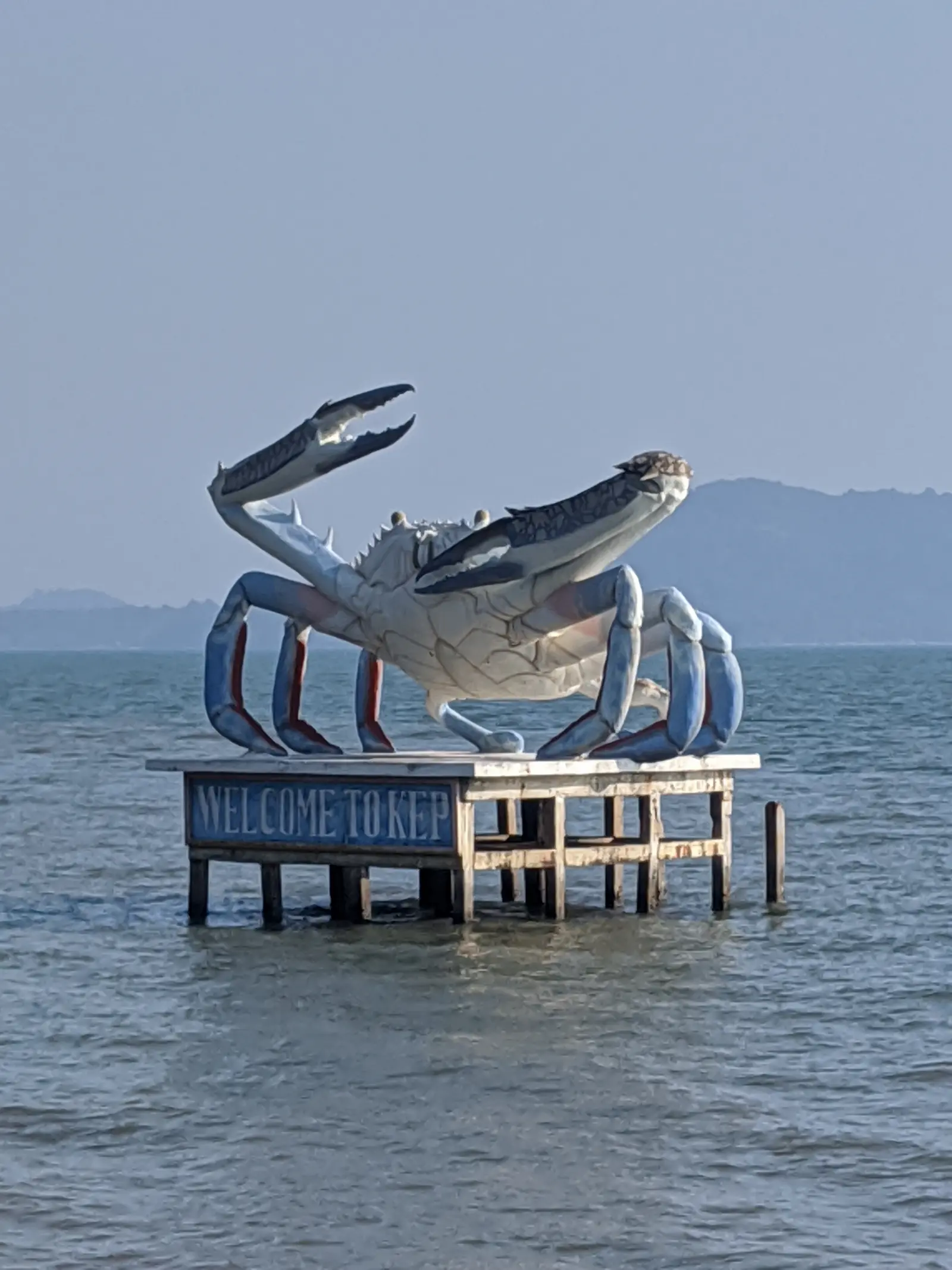 A large blue crab statue standing in the shallow sea off Kep Beach, with the Gulf of Thailand and the coastline in the background.