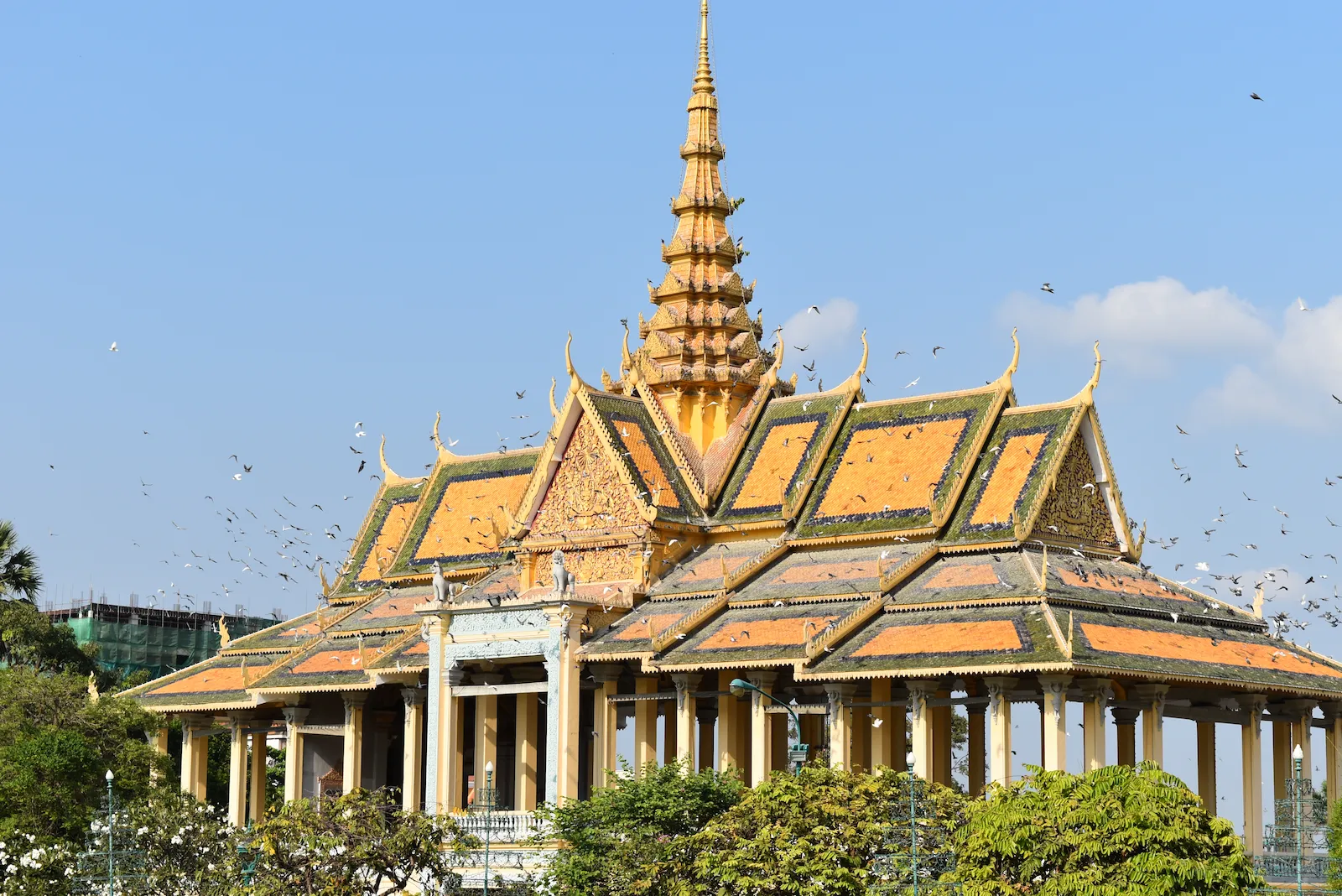 An elaborate golden pavilion with multi-layered orange roofs and a central spire at the Royal Palace in Phnom Penh, Cambodia, surrounded by a flock of flying birds against a blue sky.