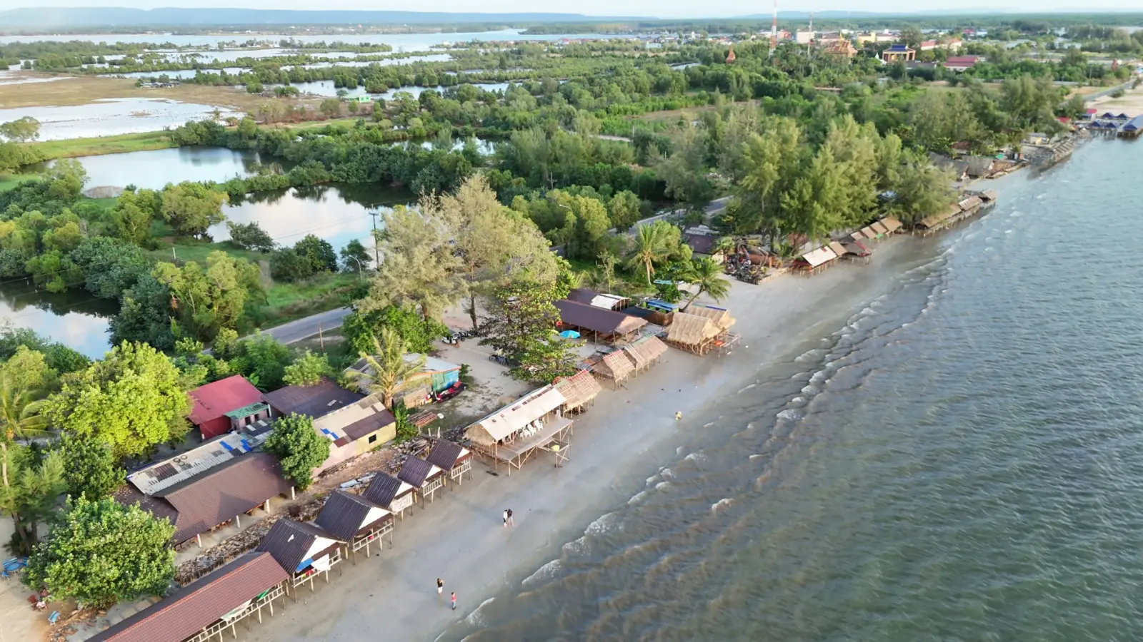 Drone shot of Pak Khlong Beach with beach huts and waves in the foreground and water filled fields in the background.
