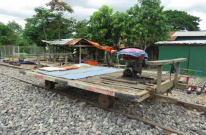 A bamboo frame sits on train wheels and on a train track, There is gravel around it with some wooden huts behind it.