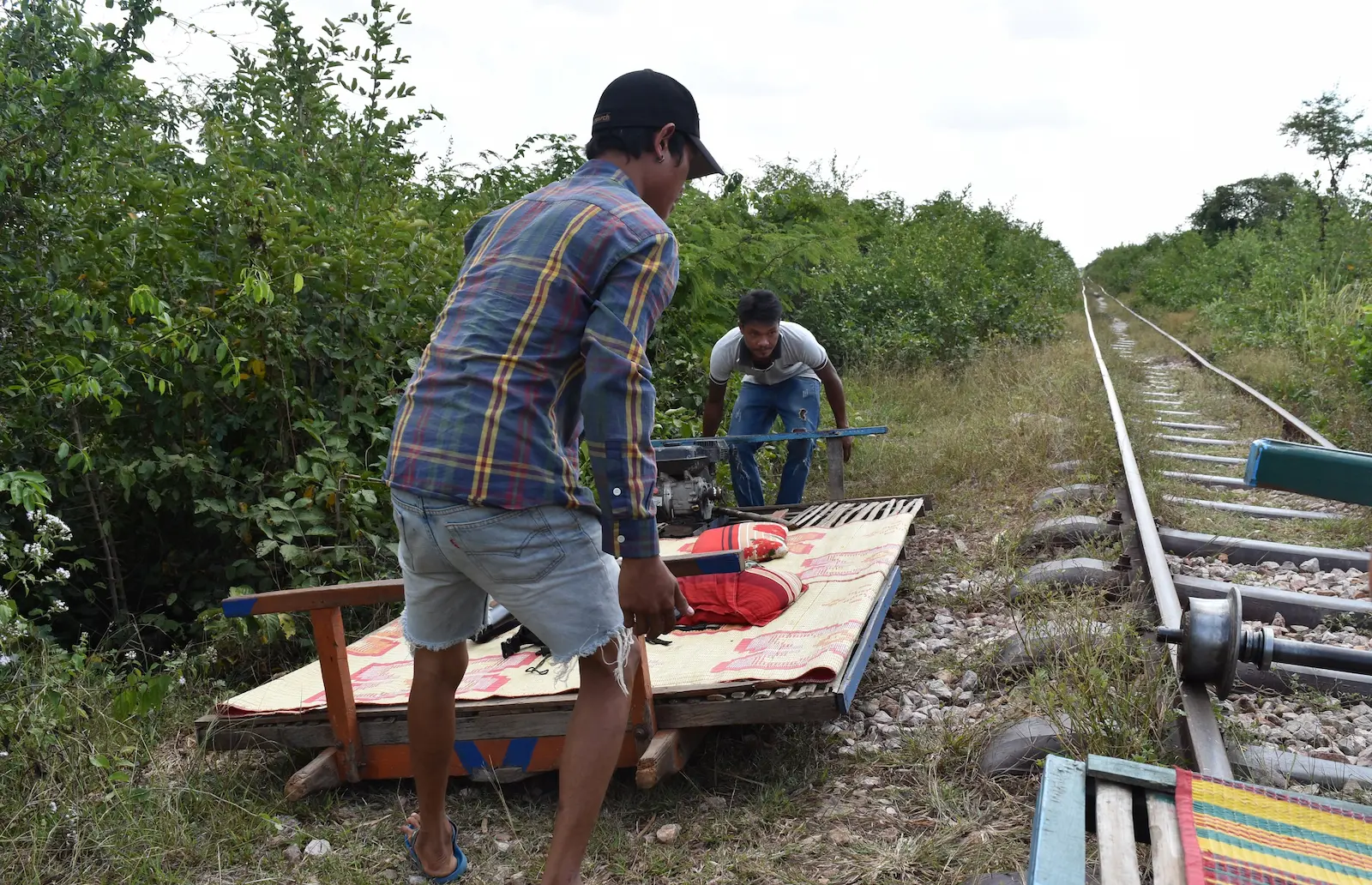 Two Cambodians lift a bamboo flatbed off its axels which are left on the train tracks next to it.