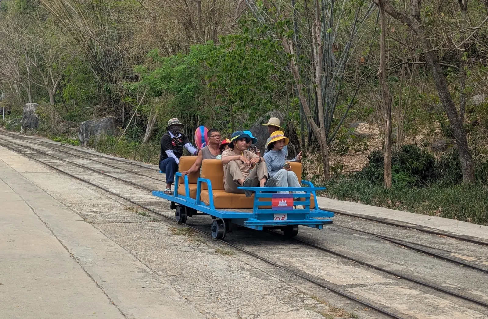 A blue steel bamboo train on the tracks with 6 people sitting on the platform