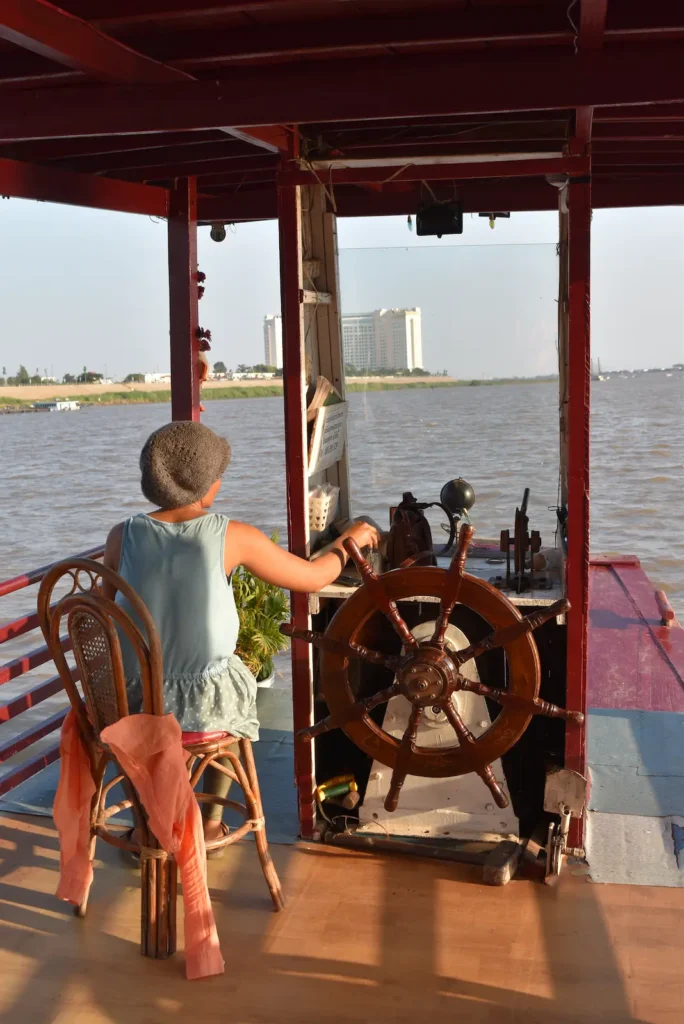 A woman is steering a boat using a large wooden wheel on the river.