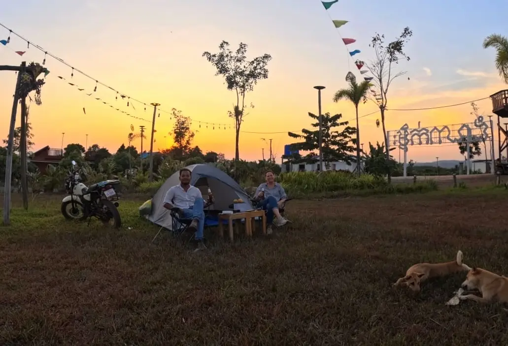 Mel and Nehru sit in front of a tent with the motorbike on their right. In front is two dogs playing and the sun is setting behind them