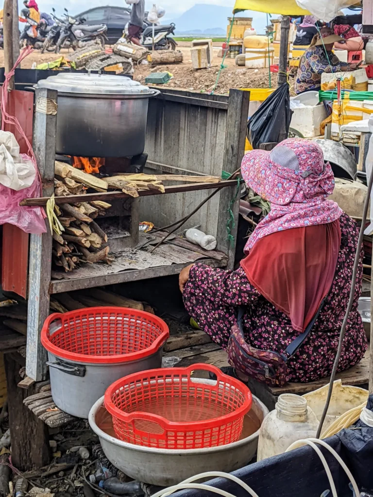A woman wearing a hat sits in front of a fire with a large pot on it. Next to her are red baskets.