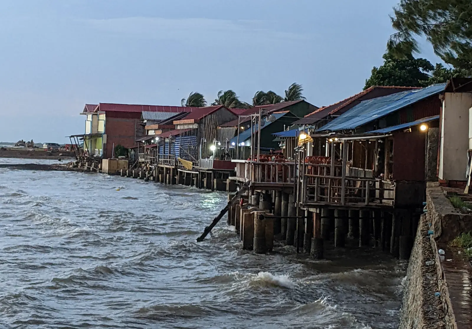 A row of wooden restaurants stand on stilts in the sea. It is dusk.