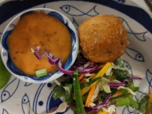A round ball which is covered in fried breadcrumbs. Next to it is a shredded salad and orange sauce in a bowl.