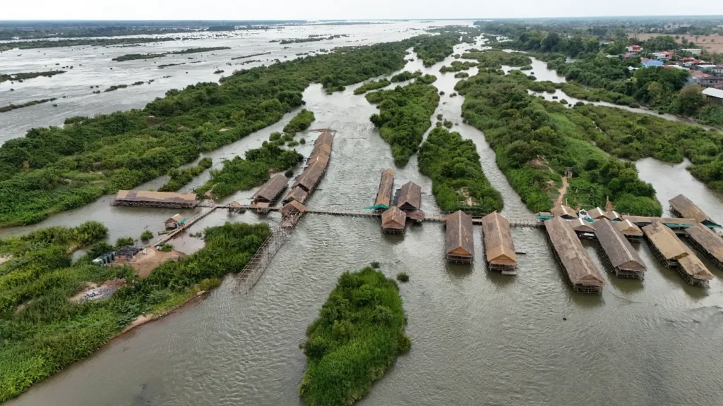 A drone shot of bamboo huts lining the water with some water bushes next to them.