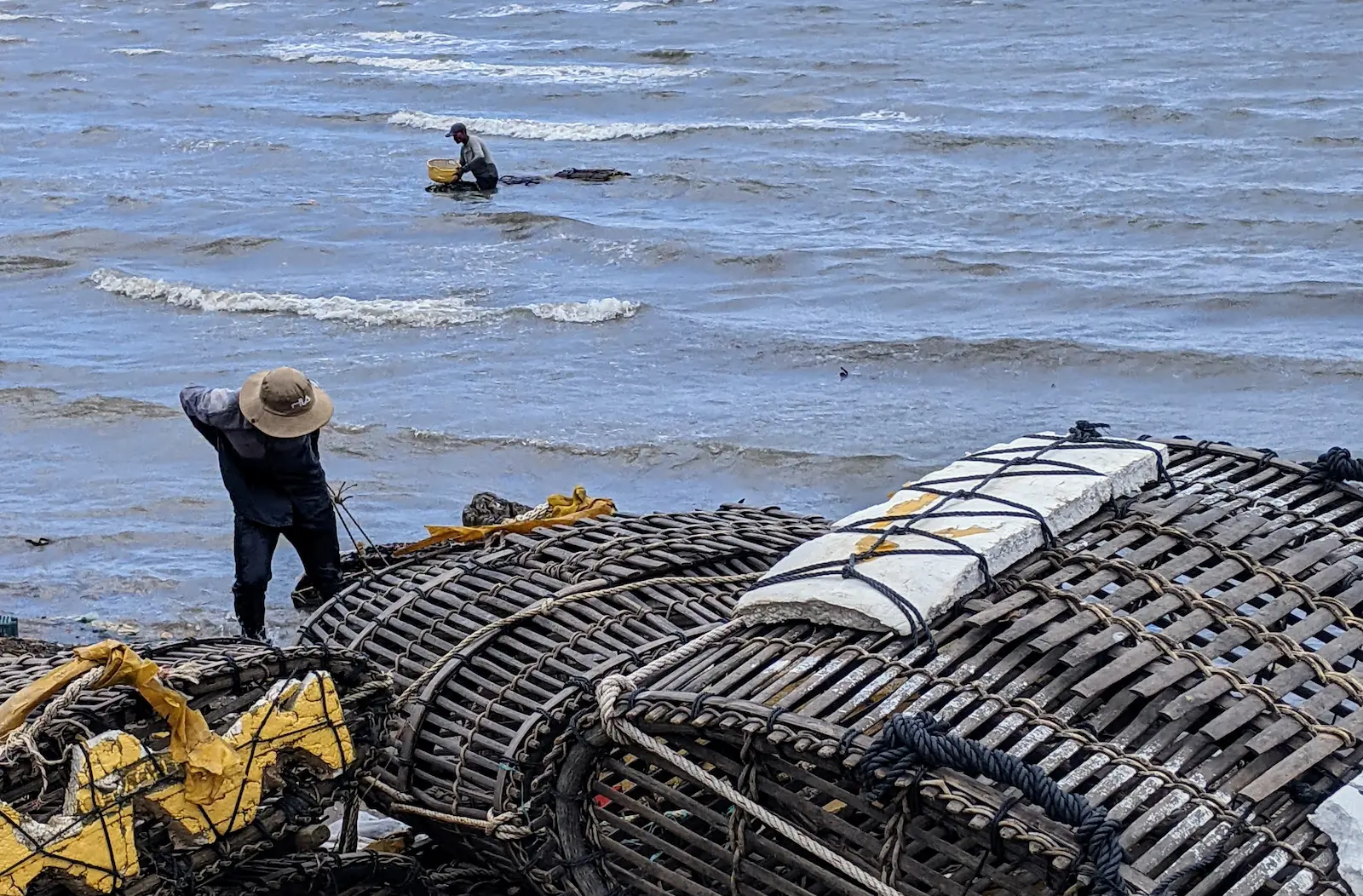 In the foreground wooden baskets are wrapped in ropes. Behind them two fishers pull in baskets from the water.