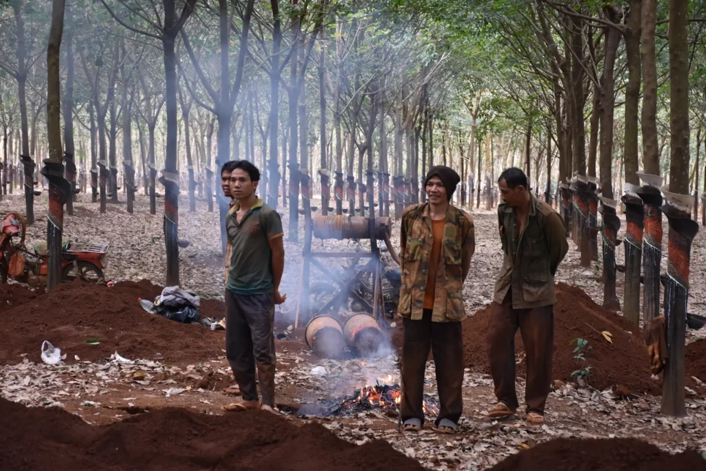 Three men covered in red dust stand next to a pit fire in a rubber plantation. Around them are mounds of dirt from mining.