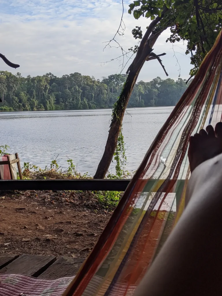 A hammock with persons leg in it is in front of a lake with a single tree.