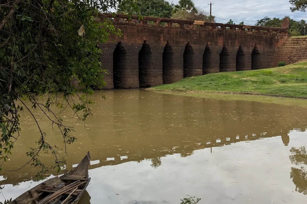 An ancient stone bridge with a river running under it. In the foregrounds is a wooden boat and a tree.