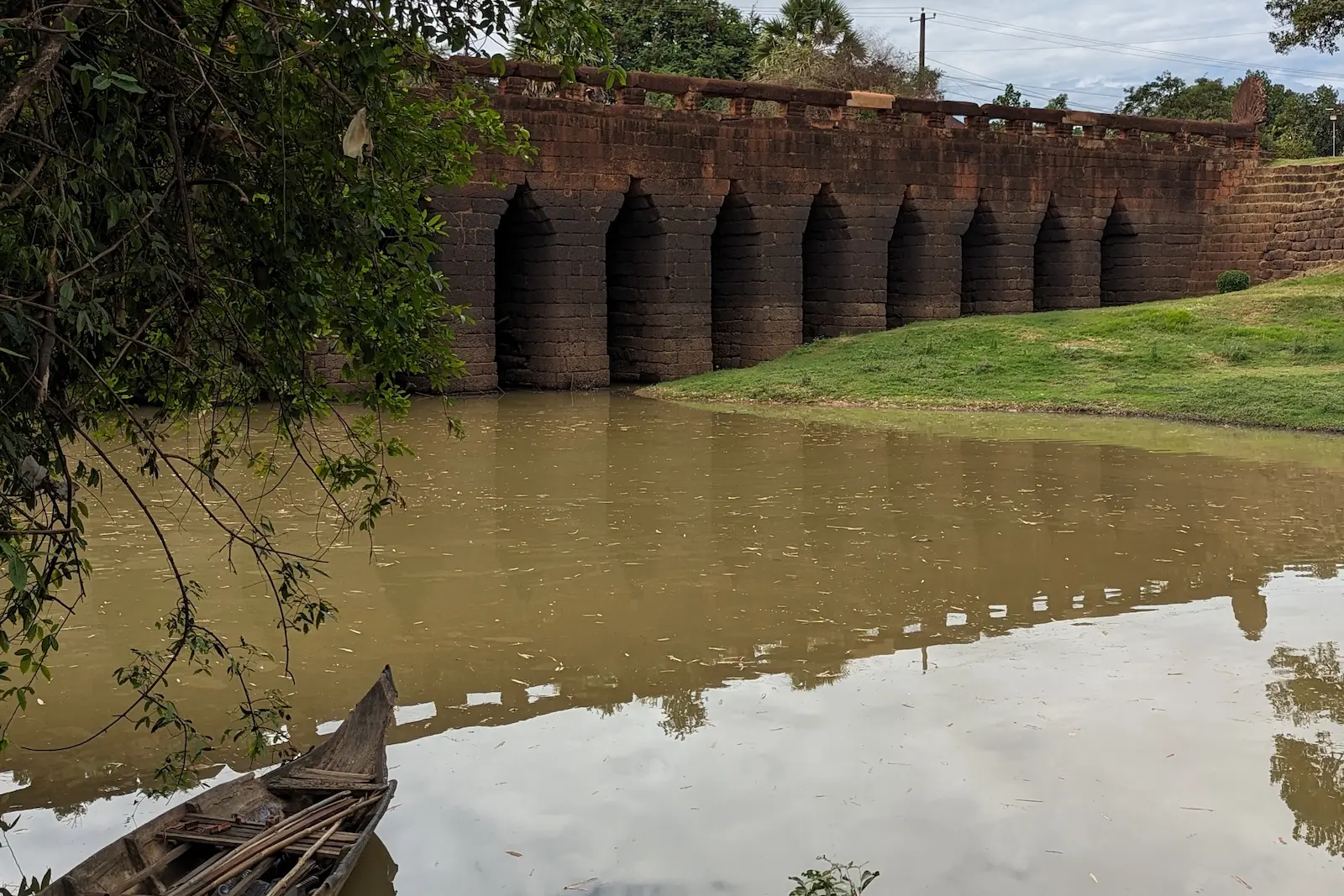 An ancient stone bridge with a river running under it. In the foregrounds is a wooden boat and a tree.