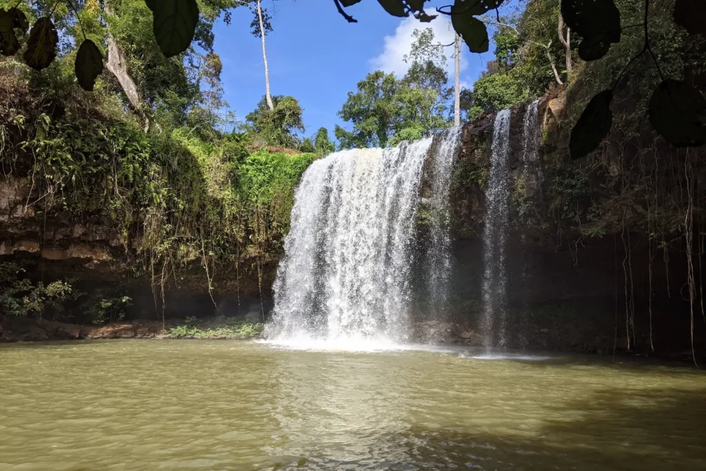 A curtain waterfall flows over rocks with lush greenery around it. A large pool of water is in front of the waterfall.