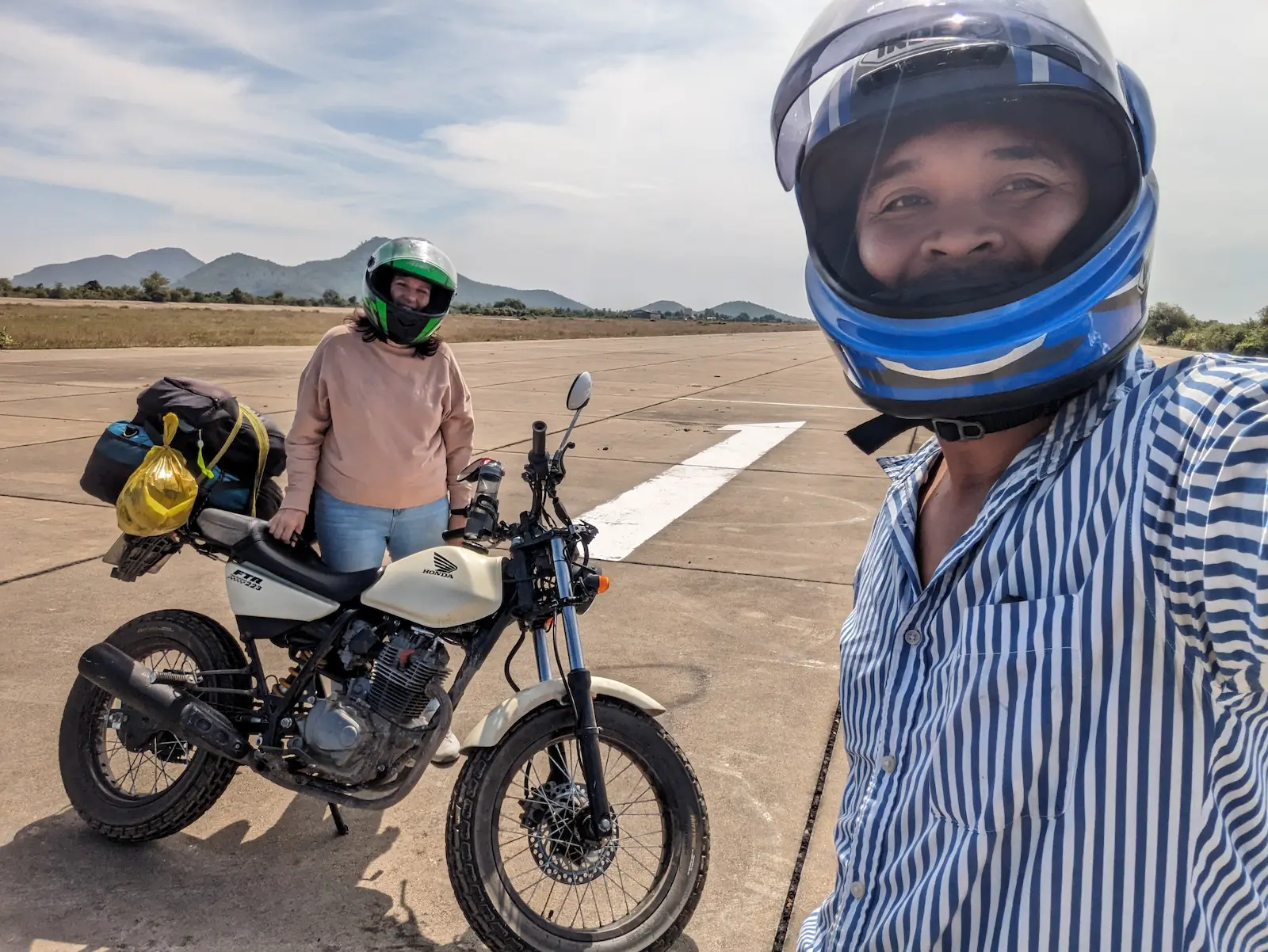 Nehru takes a selfie wearing a helmet. Mel is in teh background next to the cream FTR motorbike, also with her helmet on.
