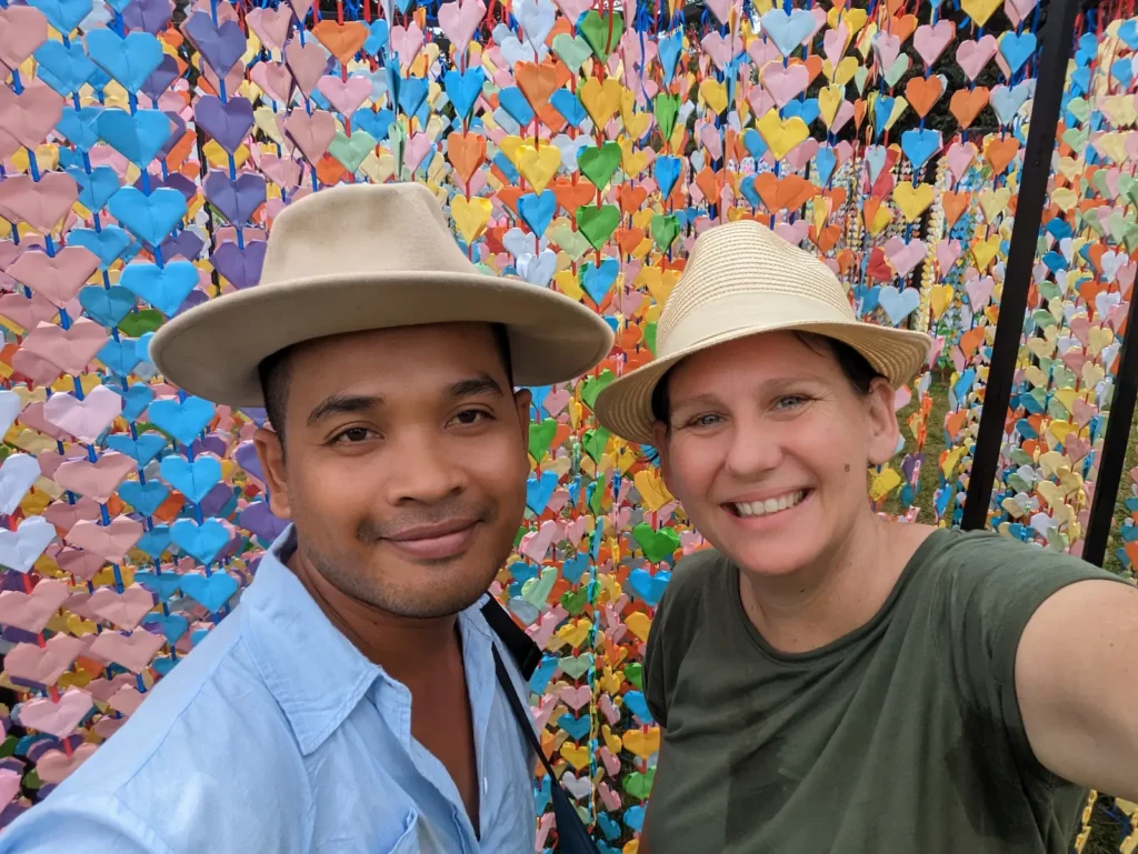 Mel and Nehru stand at Angkor Wat in front of the thousands of paper hearts they used to break the world record. They are both wearing hats and smiling.