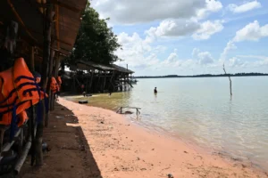 A row of huts sit on a sandy beach of West Baray reservoir. People are swimming on tyre tubes and life jackets hang on a nail.