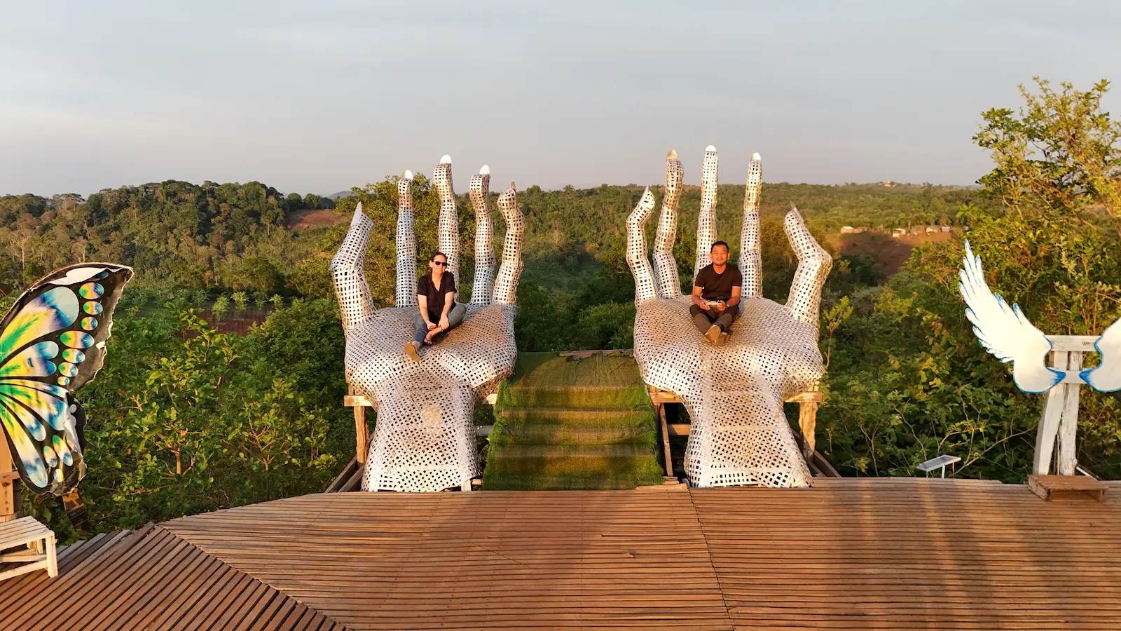 Two steel hands come out from a platform with the views of Mondulkiri behind it. Mel and Nehru sit on the giant silver hands.