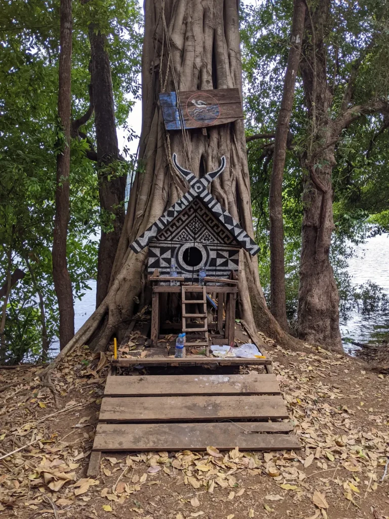 A spirit house made from wood sits in front of a tree. It is painted in black and white patterns.