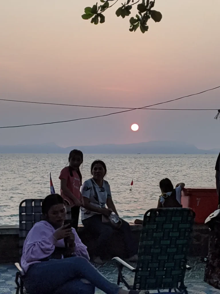 Cambodian tourists sit on chairs. Behind them the sun sets over the sea.