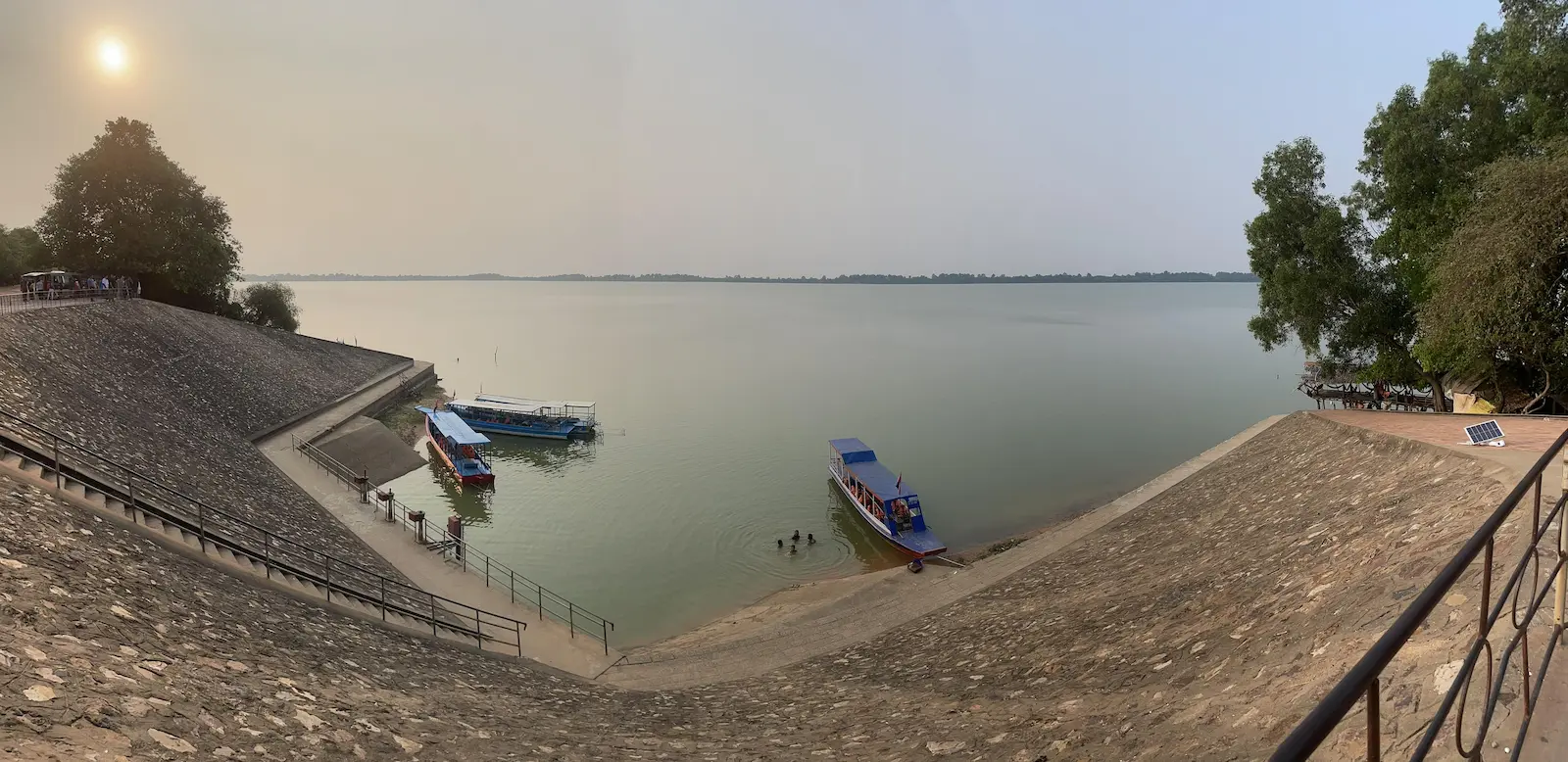 Panoramic photo taken at the edge of a spillway. In the water are some boats and children swimming. The sun is setting in the distance.