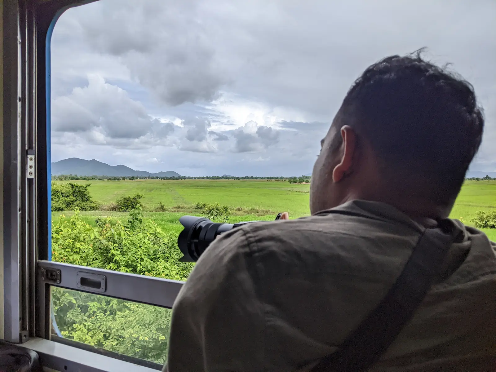 Nehru leans on the open train window with his camera. Outside is lush green fields.
