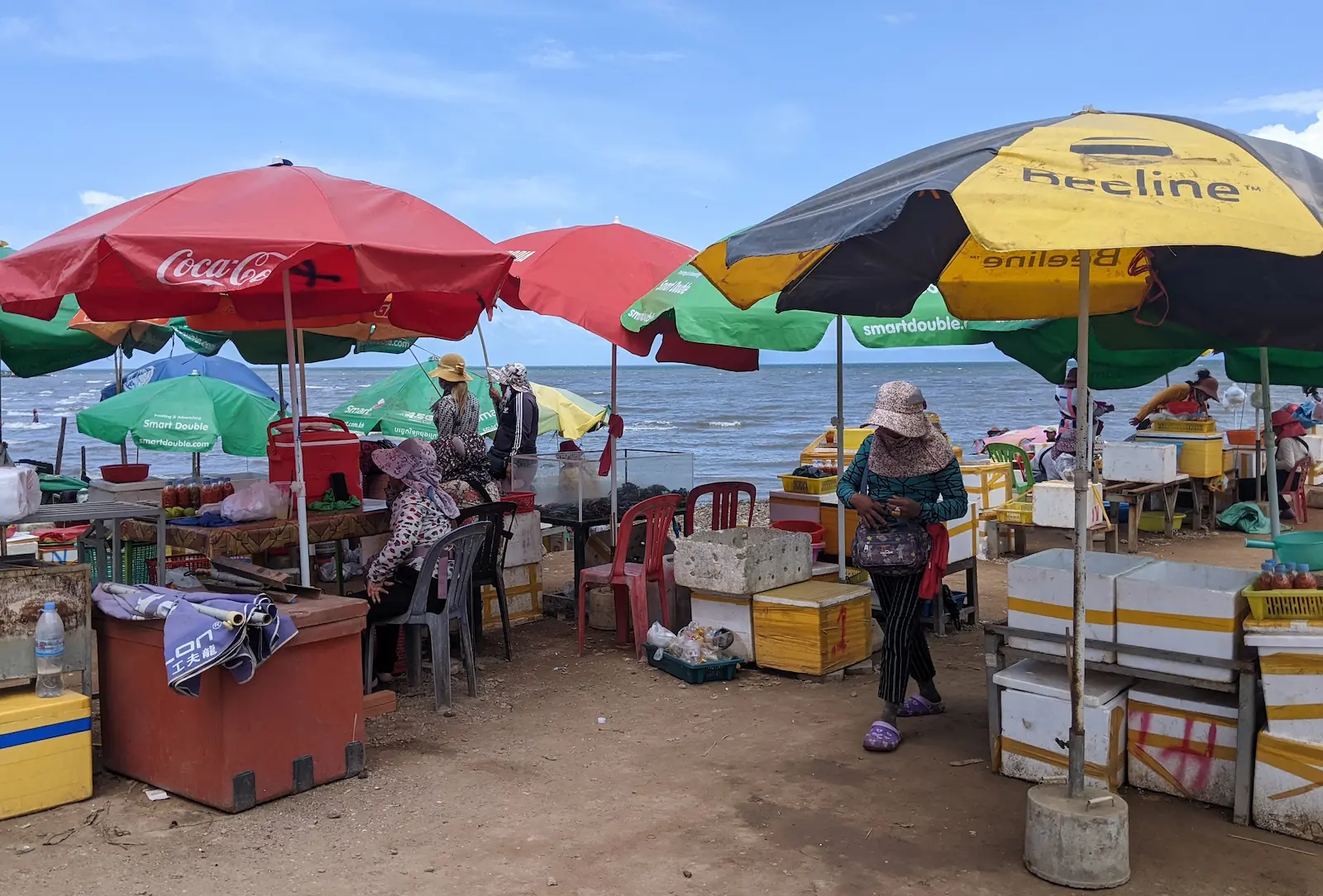 Polystyrene boxes and tables sit under umbrellas with the sea in the background. Women are selling seafood at the Kep Crab Market