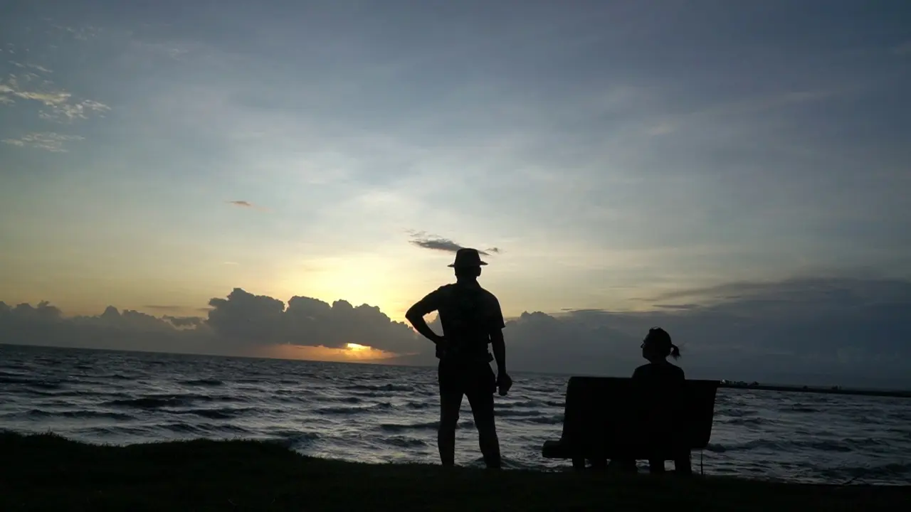 Silhouettes of two people, one standing, one sitting on the sea as the sun sets.