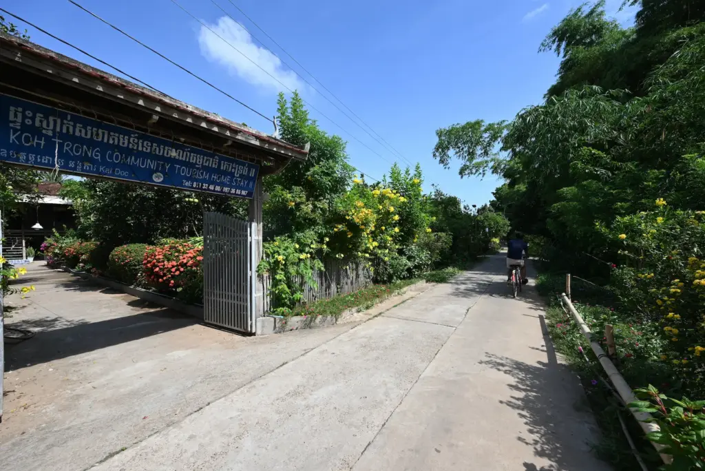 A blue sign advertises Koh Trong homestay along a concrete road. Nehru cycles his bicycle past.