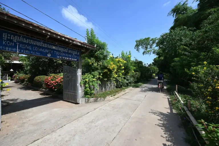 A blue sign advertises Koh Trong homestay along a concrete road. Nehru cycles his bicycle past.