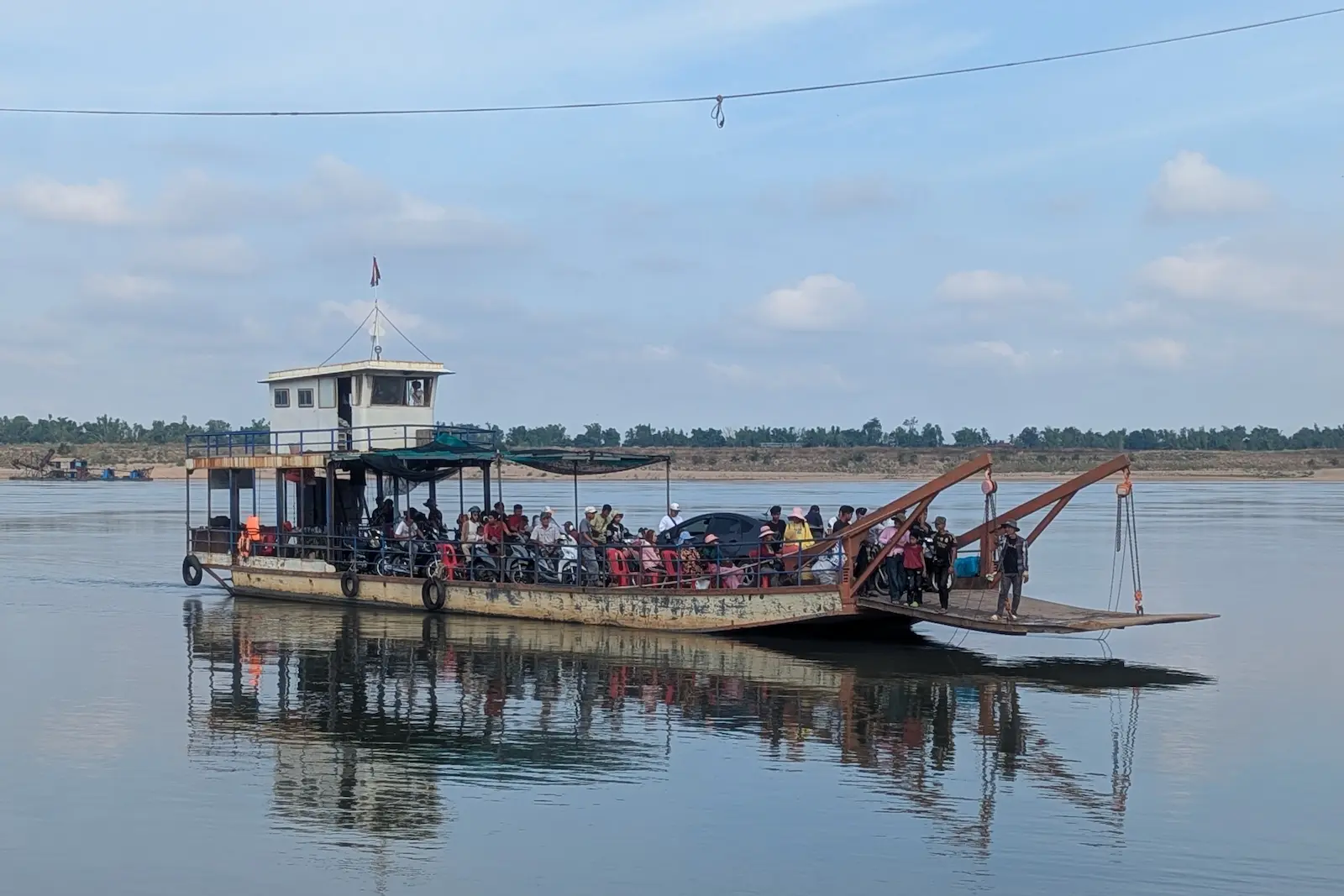 Ferry full of people, motorbikes and one car crosses a river.