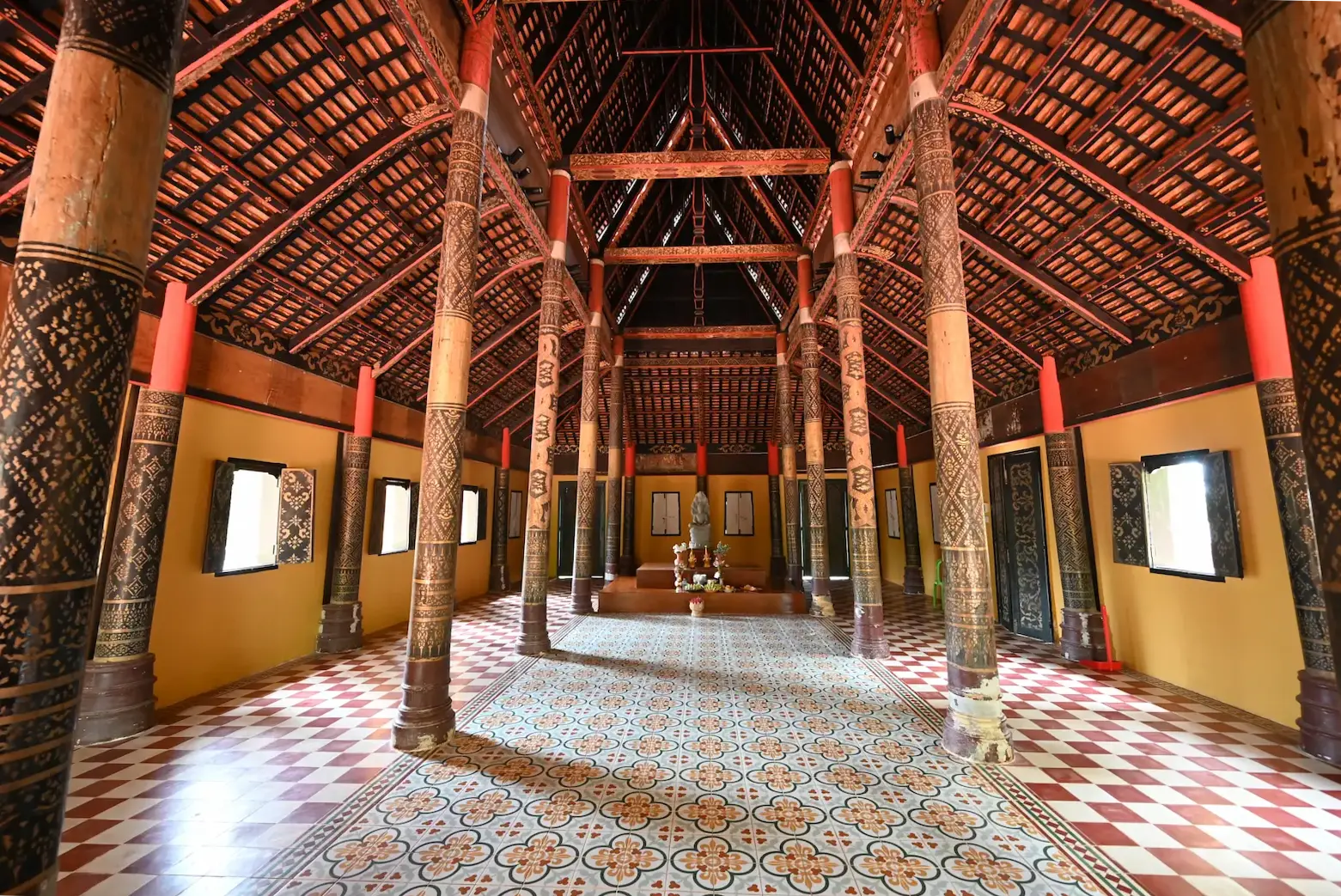 looking at the altar in a  wooden pagoda. There are large wooden pillars with stencilled designs on them and the rook trusses are carved with nagas.