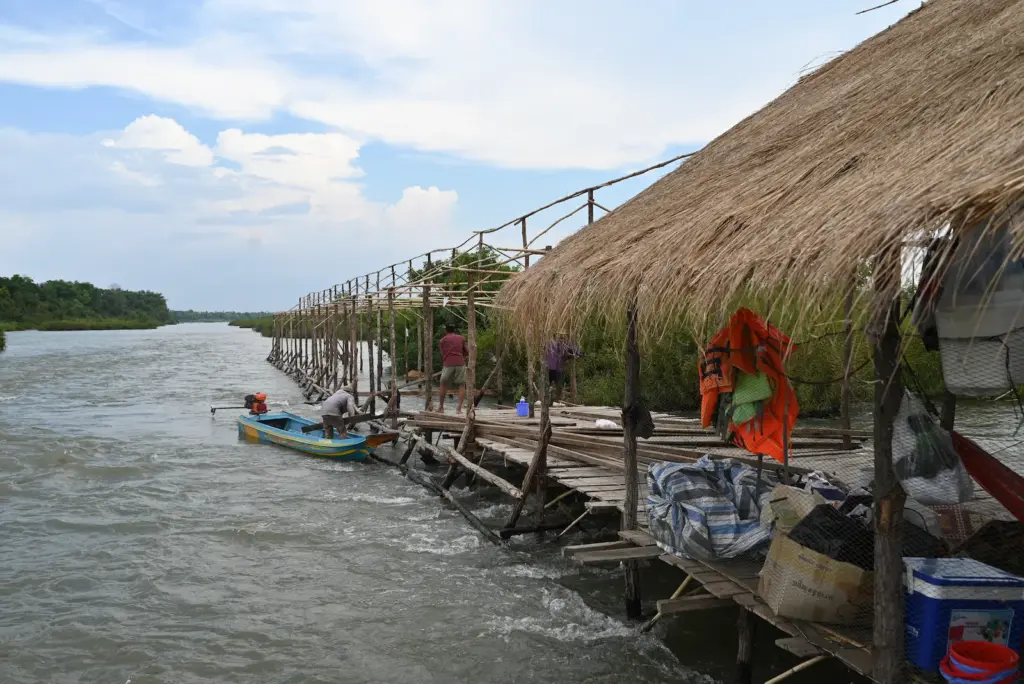Two men, one in a boat and one standing on a platform dismantle a bamboo platform over a river.