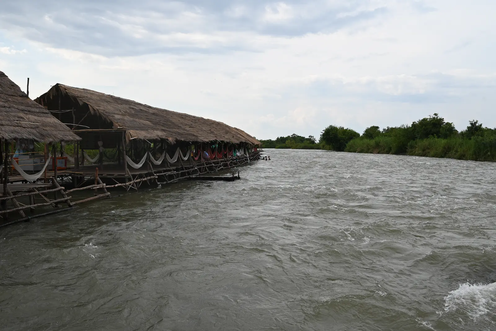 The Mekong River flows quickly next to long bamboo huts in the water. Hammocks can be seen in the huts.