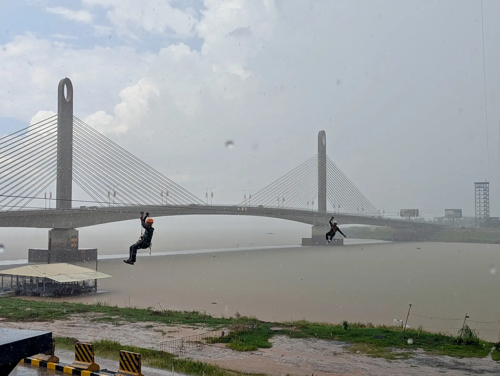 Koh Norea bridge in the background and two people ziplining in the foreground over a river.
