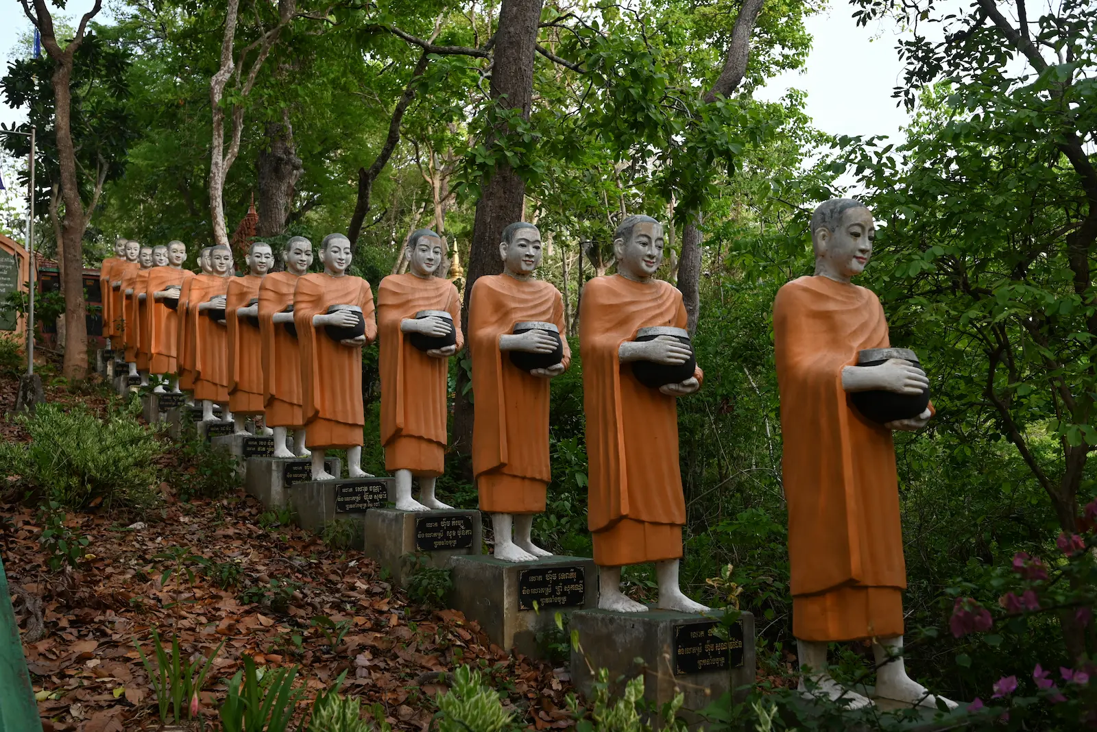 A line of monks sculptures holding alms jars are in a line coming down a hill.