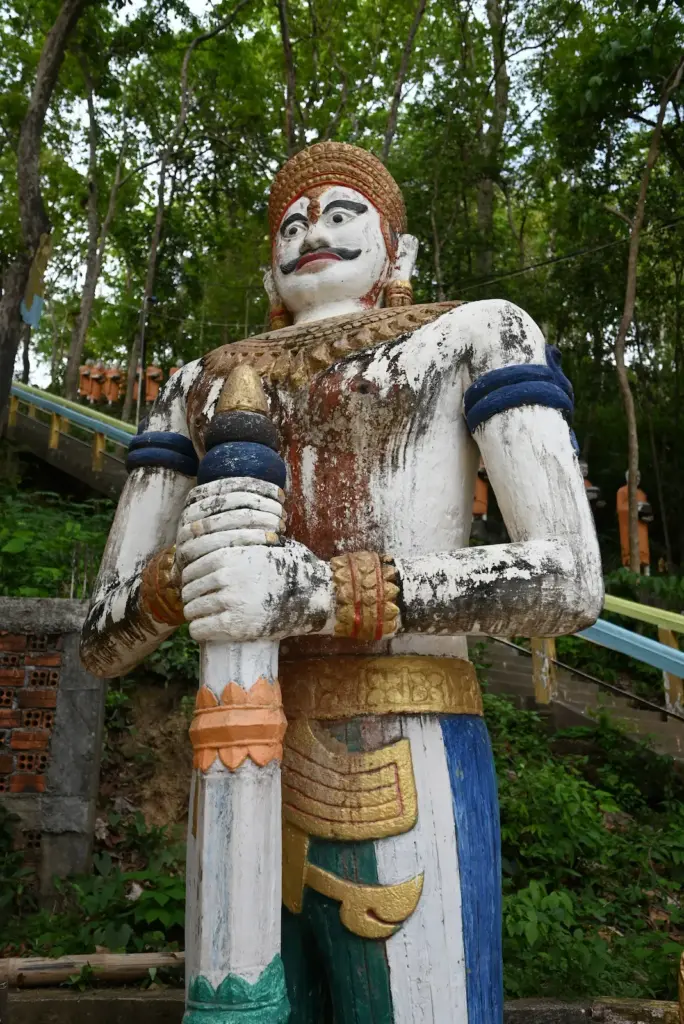 A painted guardian figure holds a staff outside stairs leading to a pagoda.