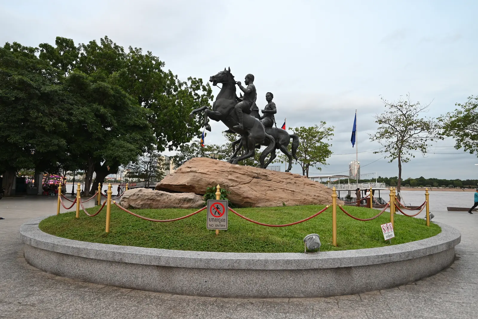 A bronze like statue of two bucking horses on a rock. The statues sit over a grassed circle next to a river.