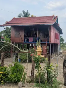 A wooden house on stilts with stairs at the front. A fence in front has a shovel leaning against it.