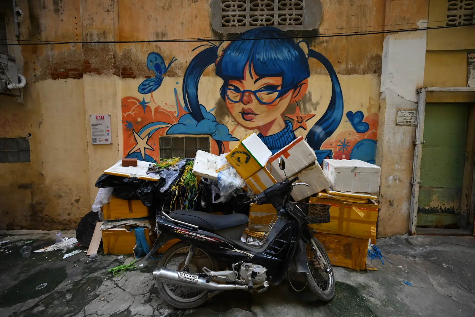 Street art of a girl with blue hair. In front is a motorbike and rubbish piled high.