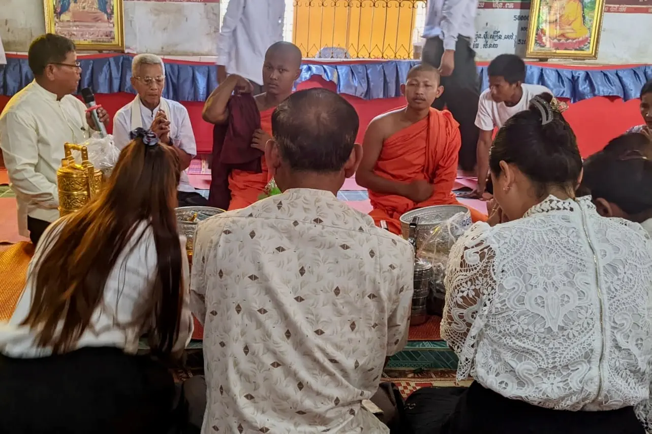 Three people wearing white shirts sit in front of two monks in a pagoda.